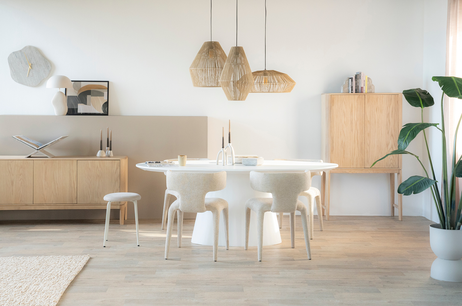 Stijlvolle eetkamer met een witte ovale eettafel van Eleonora, omringd door beige gestoffeerde stoelen en drie hangende rotan lampen. Aan de muur hangt kunst en een moderne klok, terwijl een houten dressoir en kast de ruimte completeren.